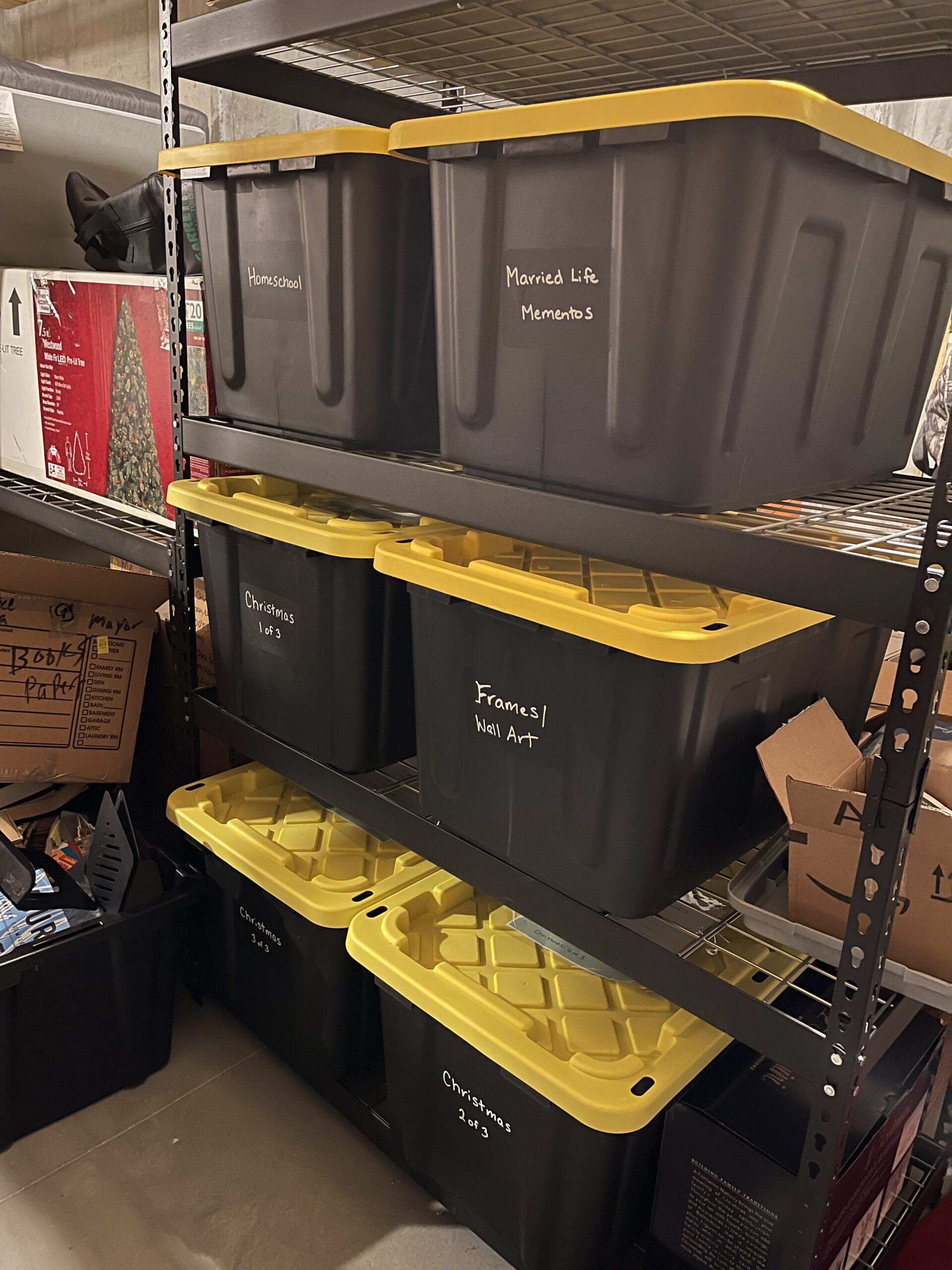 6 large plastic storage bins that are black with yellow lids, labeled with chalk stickers and chalk marker