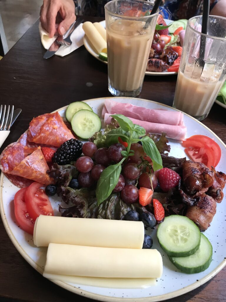 Plate with food on it: cheese rolls, cucumber slices, blueberries, grapes, bacon, sliced tomatoes, rolled cold cuts, 2 glasses of iced coffee